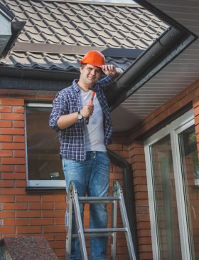 Young smiling carpenter posing with hammer on step ladder under the roof